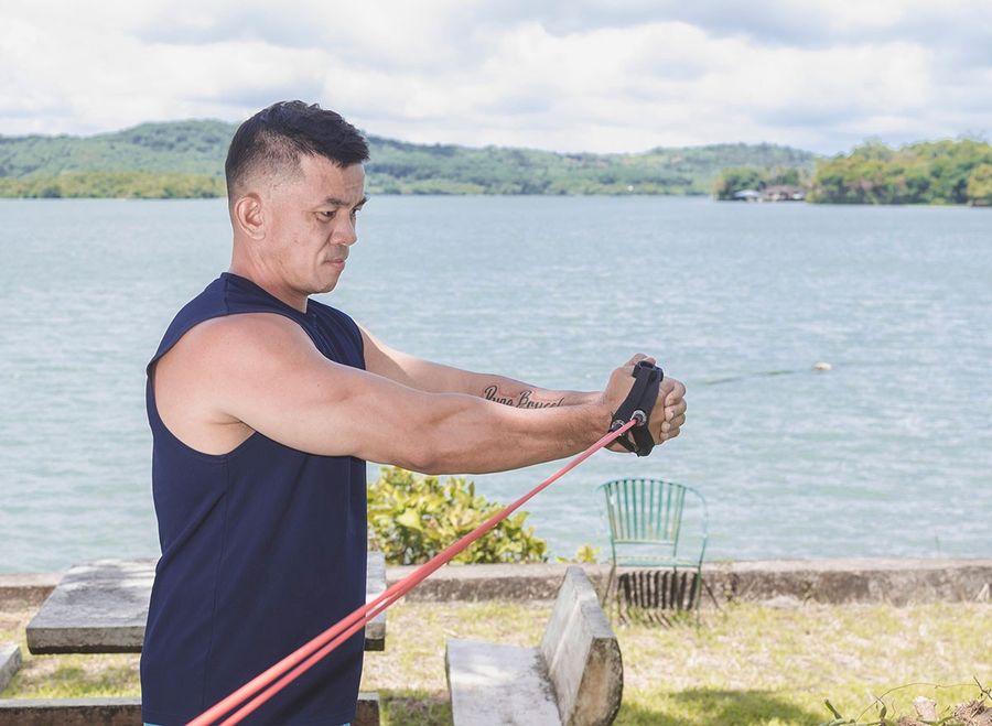 A fit middle aged man does banded low to high wood chops. A vacationer training outside at a lakeside park or resort.