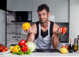 Strong healthy muscle sporty looking handsome charismatic man bodybuilder in black tank top and white apron leaning on kitchen counter with colorful vegetables, bottle of vinegar and spices on it.
