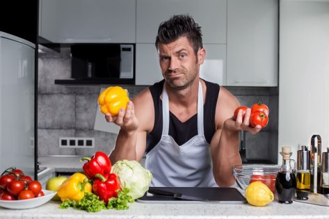 Strong healthy muscle sporty looking handsome charismatic man bodybuilder in black tank top and white apron leaning on kitchen counter with colorful vegetables, bottle of vinegar and spices on it.