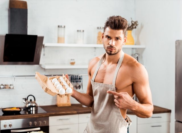 Bare-chested man in apron pointing with finger at egg carton.