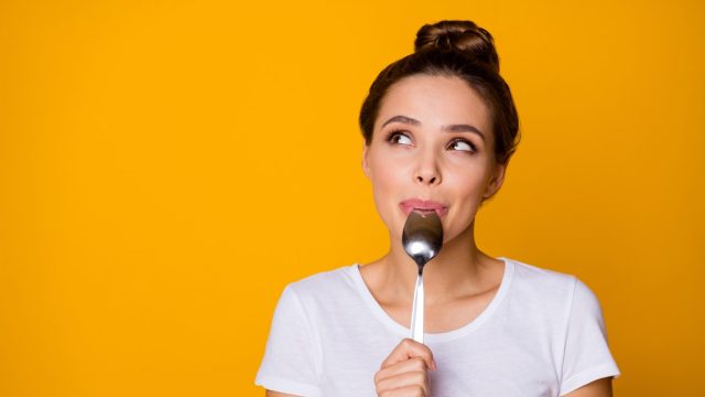 Portrait of positive inspired girl hipster lick lips spoon look copyspace feel interested about what she will eat breakfast wear white t-shirt isolated over bright shine yellow color background