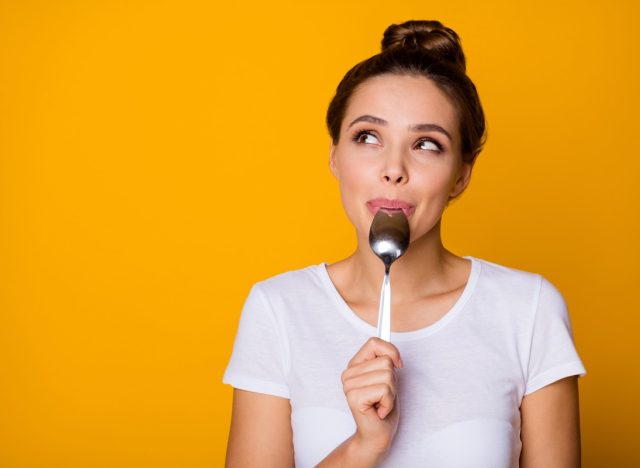 Portrait of positive inspired girl hipster lick lips spoon look copyspace feel interested about what she will eat breakfast wear white t-shirt isolated over bright shine yellow color background
