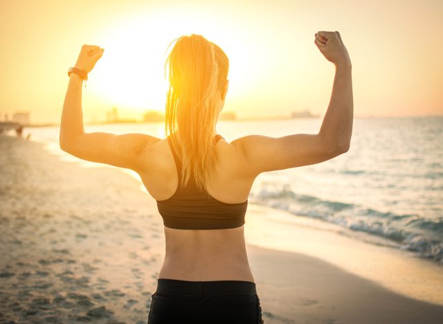 Back view of strong sporty girl showing muscles at the beach during sunset.