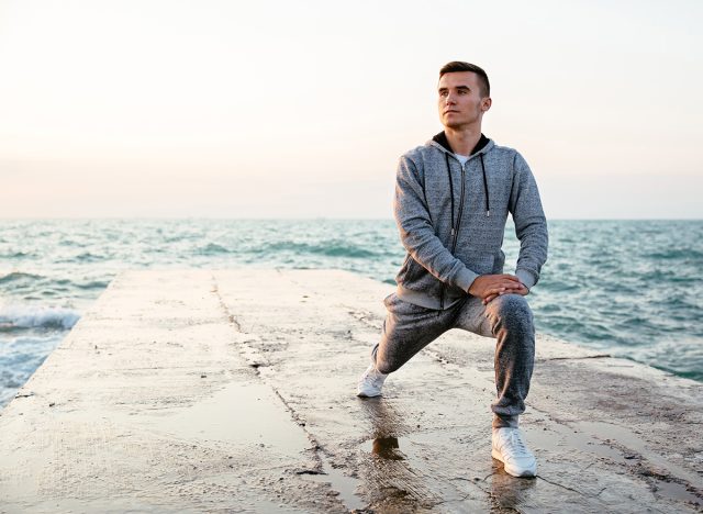 Pensive concentrated man in sportswear doing lunges, stretching, while having workout on pier, near the sea. Outdoors.