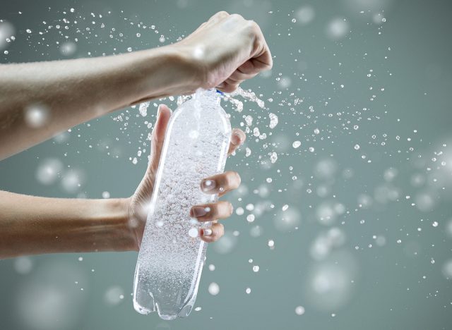 Woman's hand opening a bottle with sparkling water with splashes and lot of drops on gray background. Studio photo shooting. Concept of health lifestyle