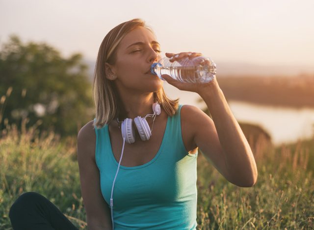 Beautiful sporty woman drinking water while sitting on exercise mat with a cityscape and river behinde her.Image is intentionally toned.