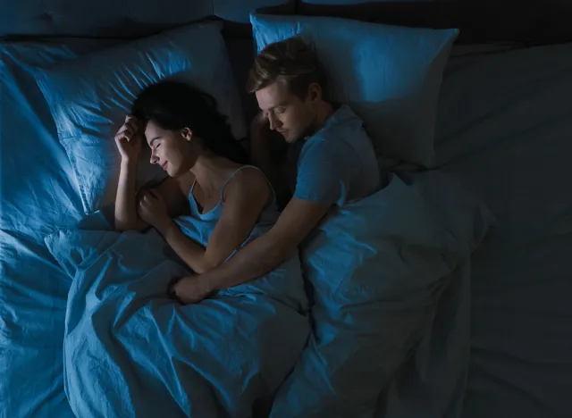 Top View Bed at Night: Attractive young Couple Sleeping Together, Holding Each other in Arms, Embracing. Blue Nightly Colors with Cold Weak Lamppost Light Shining Through the Window.
