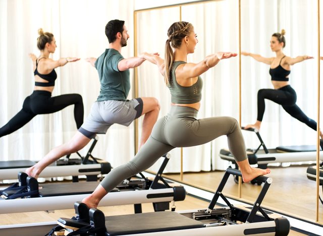 Class in a gym doing pilates standing lunges on reformer beds to stretch and tone the muscles reflected in a wall mirror