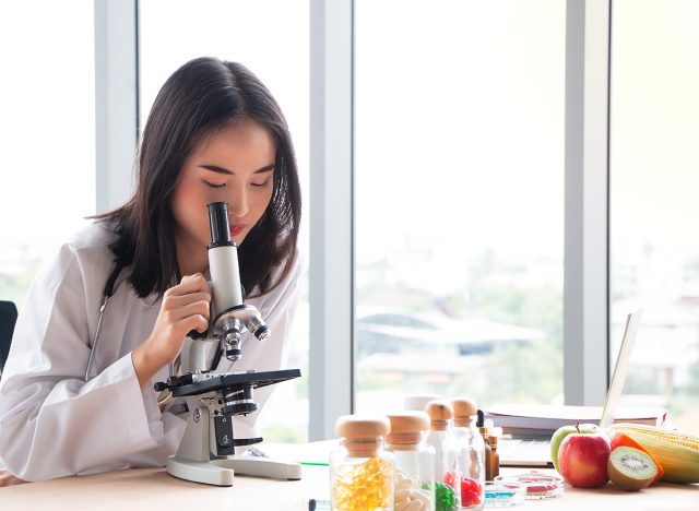 Young Asian female nutritionist doctor testing food samples with microscope on wooden table in laboratory. Fruits, vitamins and medicines on foreground. Healthy eating and diet concept with copy space