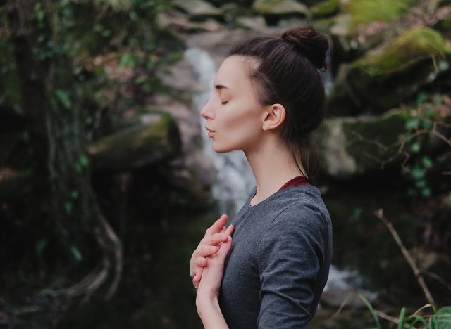 Young woman practicing breathing yoga pranayama outdoors in moss forest on background of waterfall. Unity with nature concept