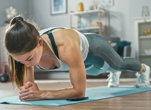 Strong Beautiful Fitness Girl in Athletic Workout Clothes is Doing a Plank Exercise While Using a Stopwatch on Her Phone. She is Training at Home in Her Living Room with Cozy Interior.