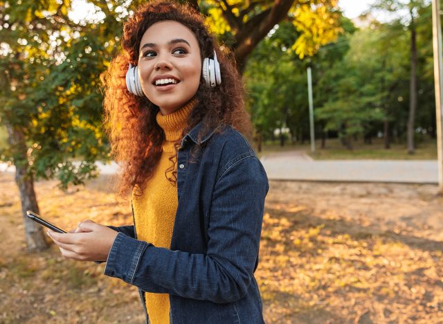 Image of a happy smiling beautiful young curly woman walking in park outdoors listening music with headphones using mobile phone.