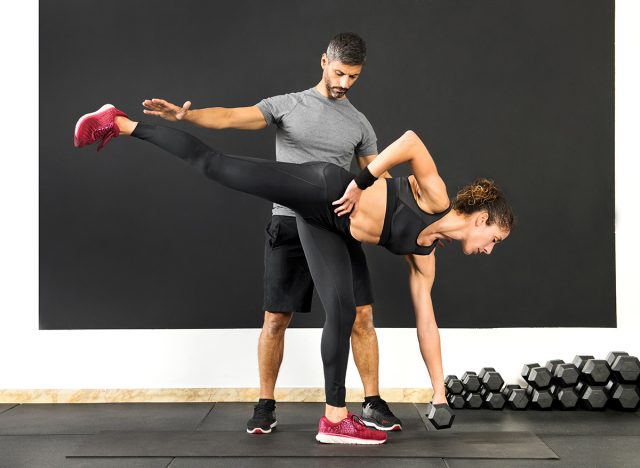 Athletic woman doing a single leg deadlift using a dumbbell weight assisted by a male personal trainer in a gym in a health and fitness concept