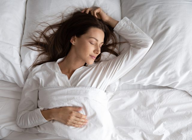 Peaceful serene beautiful young lady wear pajamas lying asleep relaxing sleeping in cozy white bed on soft pillow resting covered with blanket enjoying good healthy sleep concept, above top view