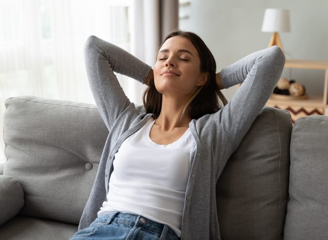 Relaxed serene pretty young woman feel fatigue lounge on comfortable sofa hands behind head rest at home, happy calm lady dream enjoy wellbeing breathing fresh air in cozy home modern living room