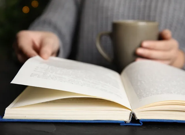 Woman with cup of beverage reading book at table, closeup