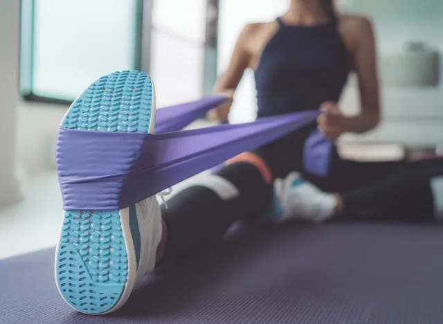 Resistance band exercise at home. Woman doing pilates workout using elastic strap pulling with arms for shoulder training on yoga mat indoors.