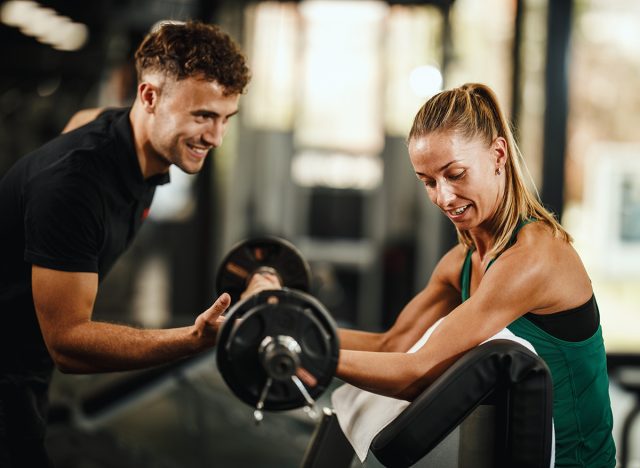 Shot of a muscular young woman in sportswear working out with personal trainer at the gym. She is pumping up her biceps muscule with heavy weight.