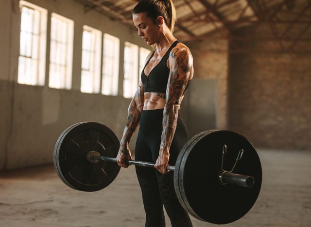 Strong woman exercising with heavy weights. Fit female doing deadlift workout with barbell in old warehouse.