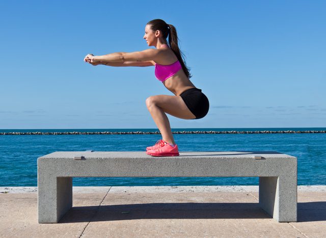 Hispanic woman doing squats on a bench by the ocean