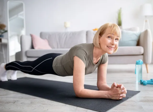 Home training concept. Smiling senior woman doing elbow plank on yoga mat in living room. Positive mature lady doing her workout routine, exercising indoors, strengthening her core muscles