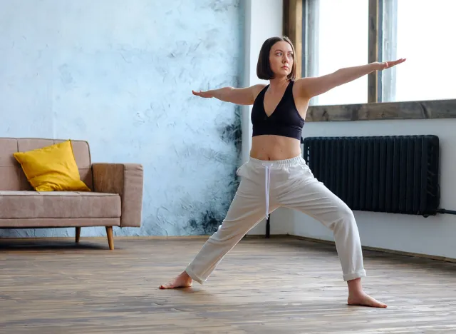 Young yogi woman practicing yoga, standing in Warrior two exercise, Virabhadrasana II pose in her living room, working out wearing sportswear bra and pants, full length. Weight loss concept .