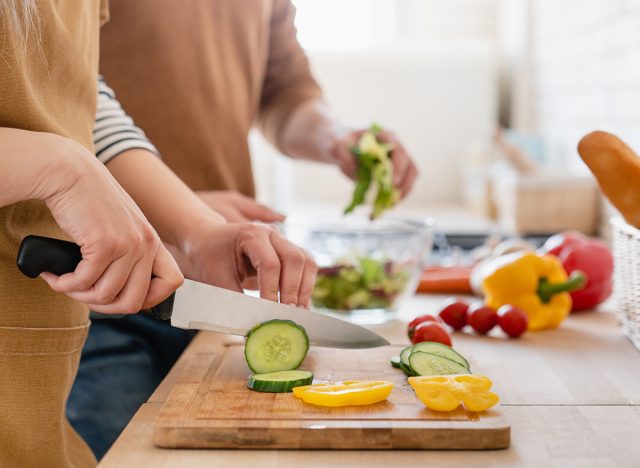 Close up cropped image of cutting board and couple cutting vegetables in the kitchen together, preparing food meal at home. Vegetarian healthy food