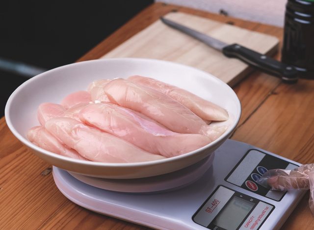 Woman in gloves puts raw chicken breasts in a plate on a digital weighing scale to prepare delicious food at home. Homemade cooking concept.