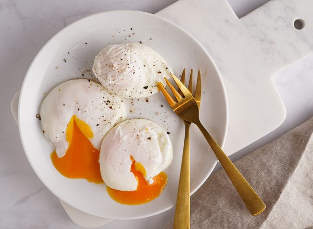 Three poached eggs with egg yolk on a white plate on a marble board and golden forks