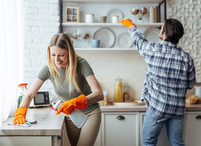 Smiling millennial european woman blonde with rubber gloves and man wipe dust on light kitchen interior, copy space. Hygiene, cleaning at home together and household chores during covid-19 outbreak