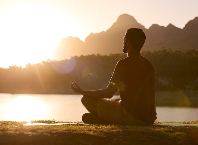 Man Meditating Doing Yoga By Lake And Mountains At Sunset