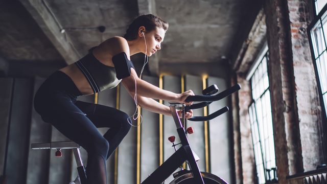 Side view of young fit female with device on arm and earphones doing cardio practice on stationary exercise bicycle in gym