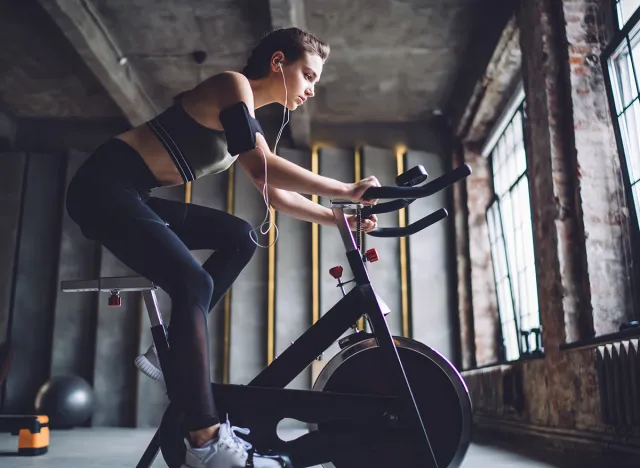 Side view of young fit female with device on arm and earphones doing cardio practice on stationary exercise bicycle in gym