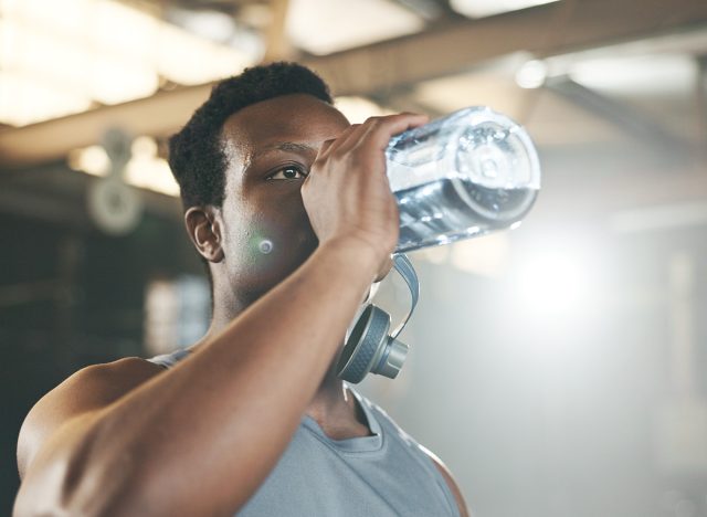 Black man at gym, water bottle and relax to hydrate in muscle development, strong body and fitness. Commitment, motivation and bodybuilder with drink in workout challenge for health and wellness.