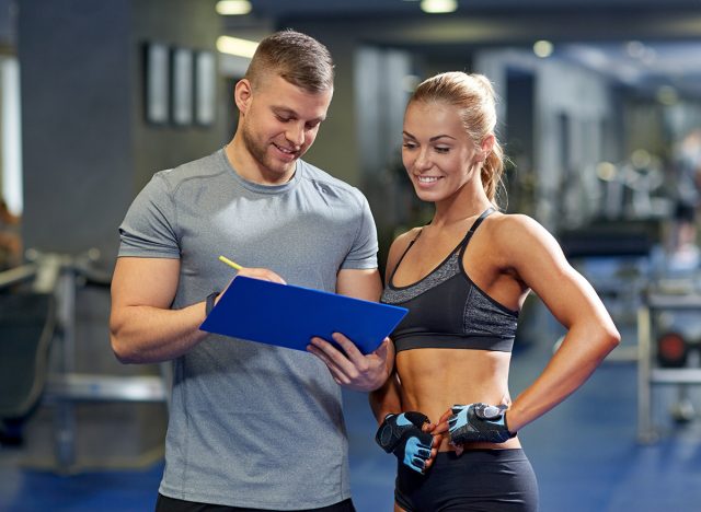 fitness, sport, exercising and diet concept - smiling young woman and personal trainer with clipboard writing exercise plan in gym