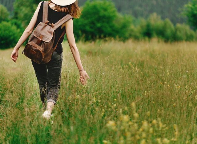 stylish hipster woman walking in grass and holding in hand herb wildflowers in summer mountains, travel concept, peaceful relaxing moment. happy earth day