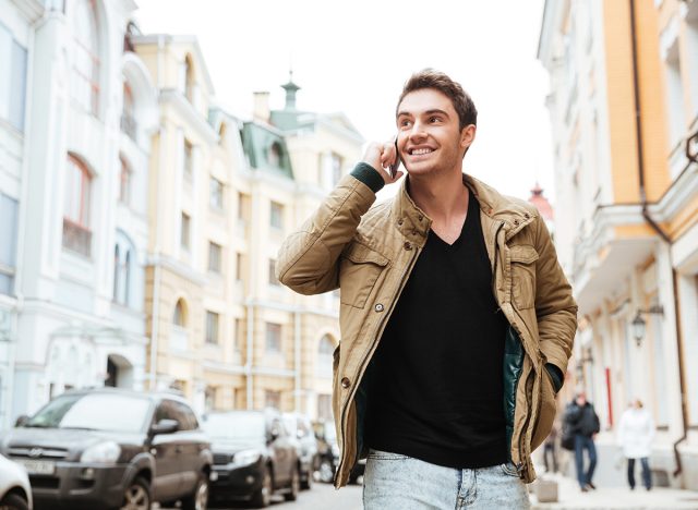 Image of happy young man walking on the street and looking aside while talking by his phone.