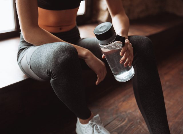 Cropped photo of attractive fitness woman sitting in gym and holding bottle of water.