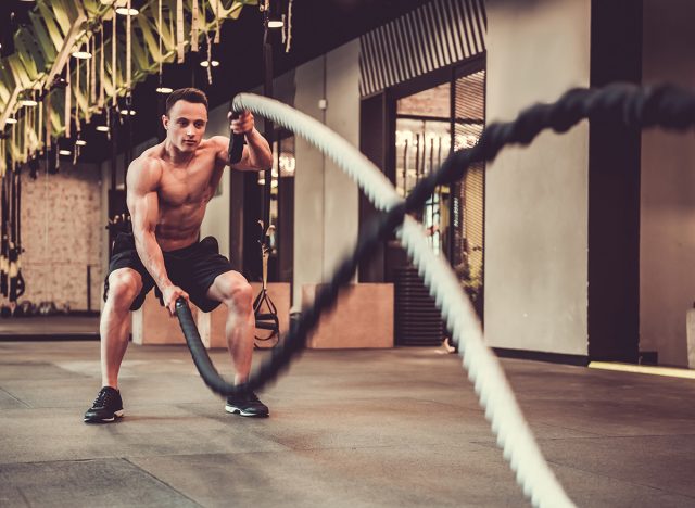 Handsome muscular man is doing battle rope exercise while working out in gym