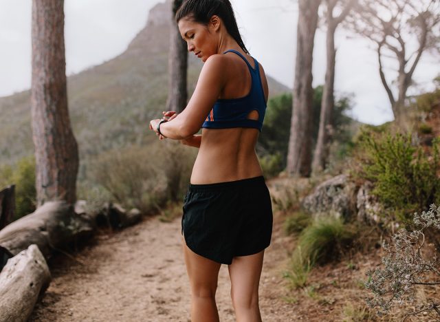 Female runner checking fitness progress on her smart watch. Asian woman using fitness app to monitor workout performance, while walking through mountain trail.
