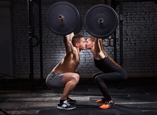 Young beautiful sportive woman and man kissing and lifting a dumbbell from squats against brick wall in the gym.