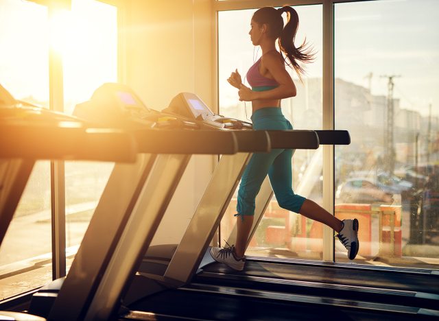 Young healthy athletic woman running on a treadmill near the sunny window in the gym and listening music.