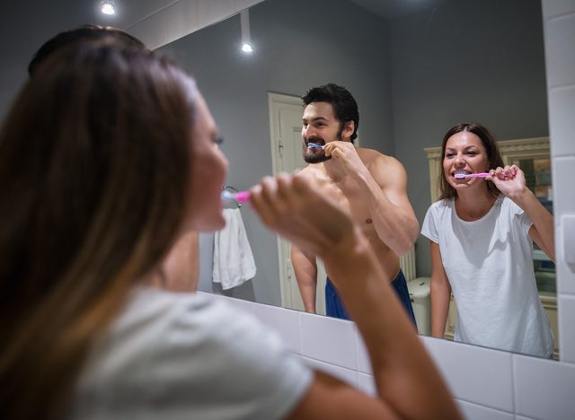 Cheerful attractive couple in love having tooth brushing together in front of mirror in the morning at bathroom.