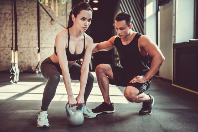 Woman is doing exercises with a kettlebell while working out with a personal trainer in gym.