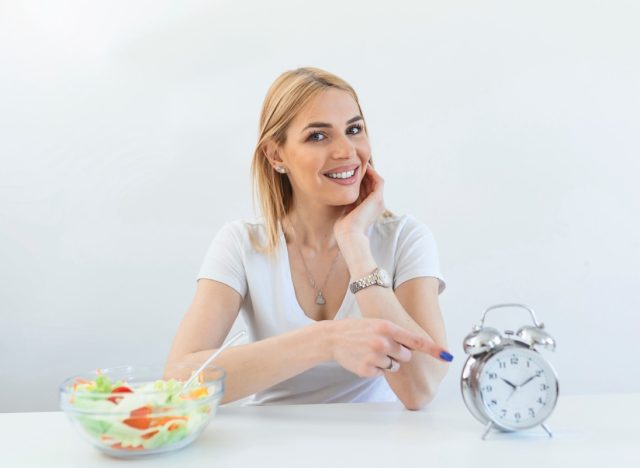 Dieting woman pointing on a clock wit salad on her side.