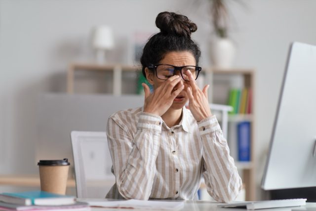 Female office worker in glasses rubbing tired eyes, exhausted from overworking, sitting at workplace in office.