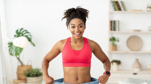 Happy African Woman After Slimming Showing Her Old Oversize Pants Standing Indoors, Smiling To Camera