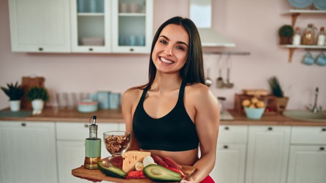 Attractive sporty woman standing on kitchen with healhy food full of protein in hand, smiling and looking at camera.