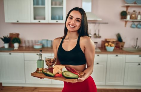 Attractive sporty woman standing on kitchen with healhy food full of protein in hand, smiling and looking at camera.