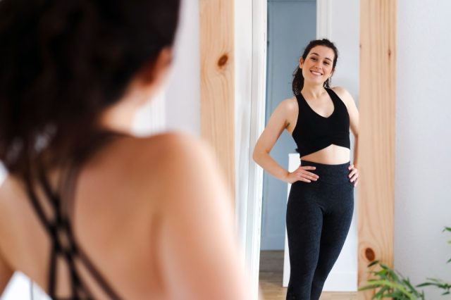 Young happy beautiful woman looking in the mirror satisfied with the results of her training.